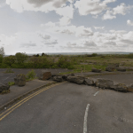 A wide view of a disused car park and overgrown land at the former Penplas supermarket site, with large rocks blocking vehicle access and open fields stretching into the distance.