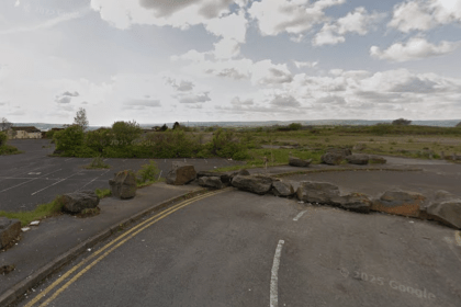 A wide view of a disused car park and overgrown land at the former Penplas supermarket site, with large rocks blocking vehicle access and open fields stretching into the distance.
