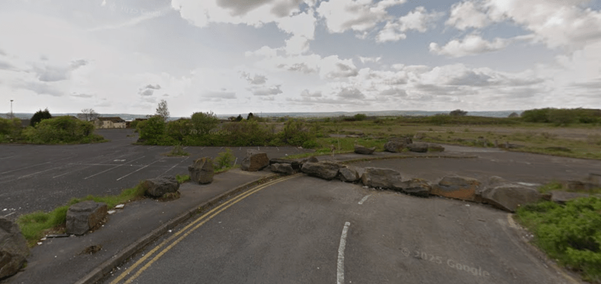 A wide view of a disused car park and overgrown land at the former Penplas supermarket site, with large rocks blocking vehicle access and open fields stretching into the distance.