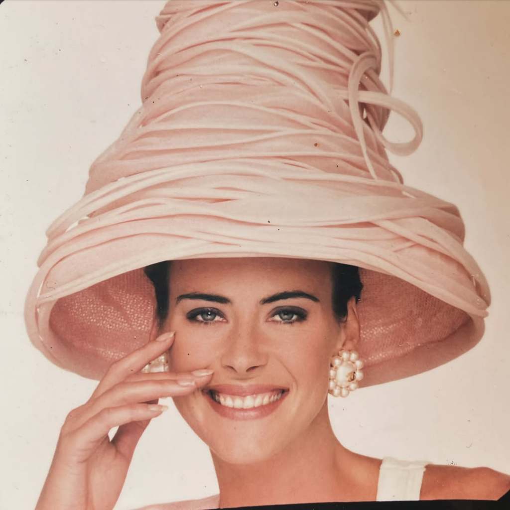 A 1980s fashion photograph of a young Annabel Schofield smiling and wearing a large, sculptural pale pink wide-brimmed hat and pearl cluster earrings, with her hand raised playfully to her face.