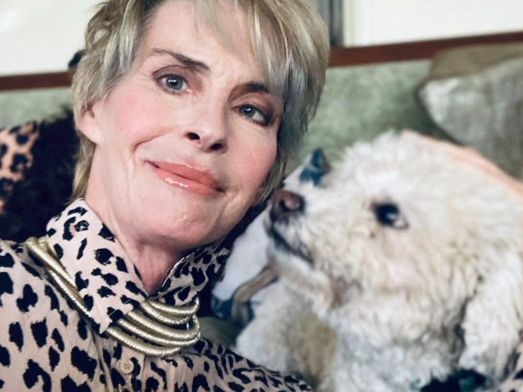 Annabel Schofield smiling in a leopard-print top and gold necklace, sitting beside a small fluffy white dog on a sofa in what appears to be a living room.