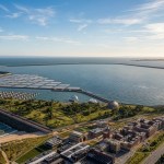 Wide aerial artist’s impression of a tidal lagoon in Swansea Bay, with a curved sea wall enclosing water, solar installations, and nearby coastal developments along the shoreline.