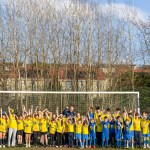A wide-angle shot of a large group of young football players in yellow and blue kits cheering in front of a goal at Trallwn Fields, flanked by club and council officials. Bare winter trees and residential houses are visible in the background.