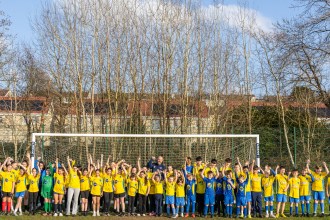 A wide-angle shot of a large group of young football players in yellow and blue kits cheering in front of a goal at Trallwn Fields, flanked by club and council officials. Bare winter trees and residential houses are visible in the background.