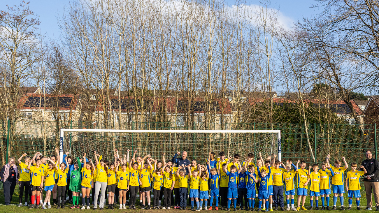 A wide-angle shot of a large group of young football players in yellow and blue kits cheering in front of a goal at Trallwn Fields, flanked by club and council officials. Bare winter trees and residential houses are visible in the background.