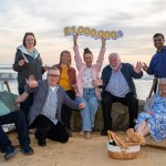 Lottery winners celebrating on a beach with arms raised, holding a £1 million sign after a EuroMillions win.