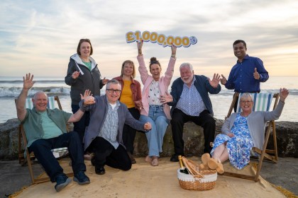 Lottery winners celebrating on a beach with arms raised, holding a £1 million sign after a EuroMillions win.