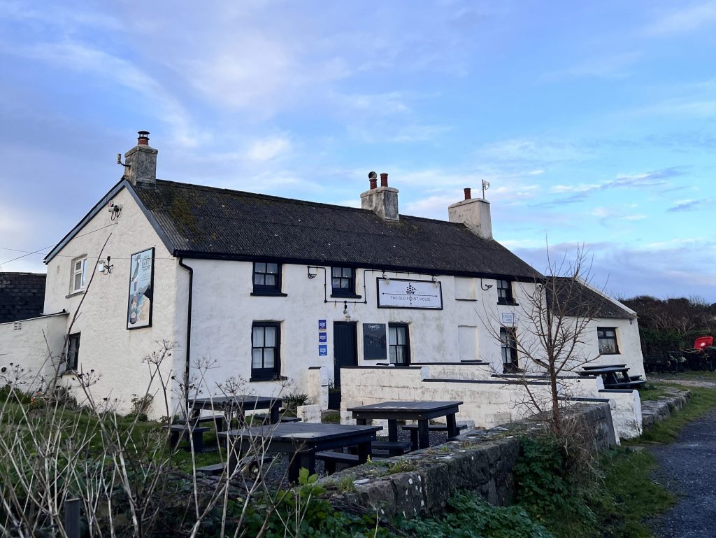 Exterior view of a traditional white coastal pub building with outdoor seating, set under a wide sky in a rural location.