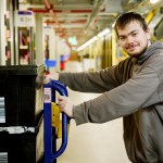 A young man in a grey hoodie stands beside a warehouse cart stacked with storage bins inside Amazon’s Swansea fulfilment centre.