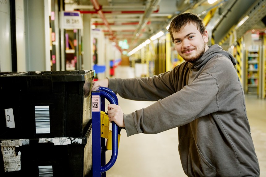 A young man in a grey hoodie stands beside a warehouse cart stacked with storage bins inside Amazon’s Swansea fulfilment centre.