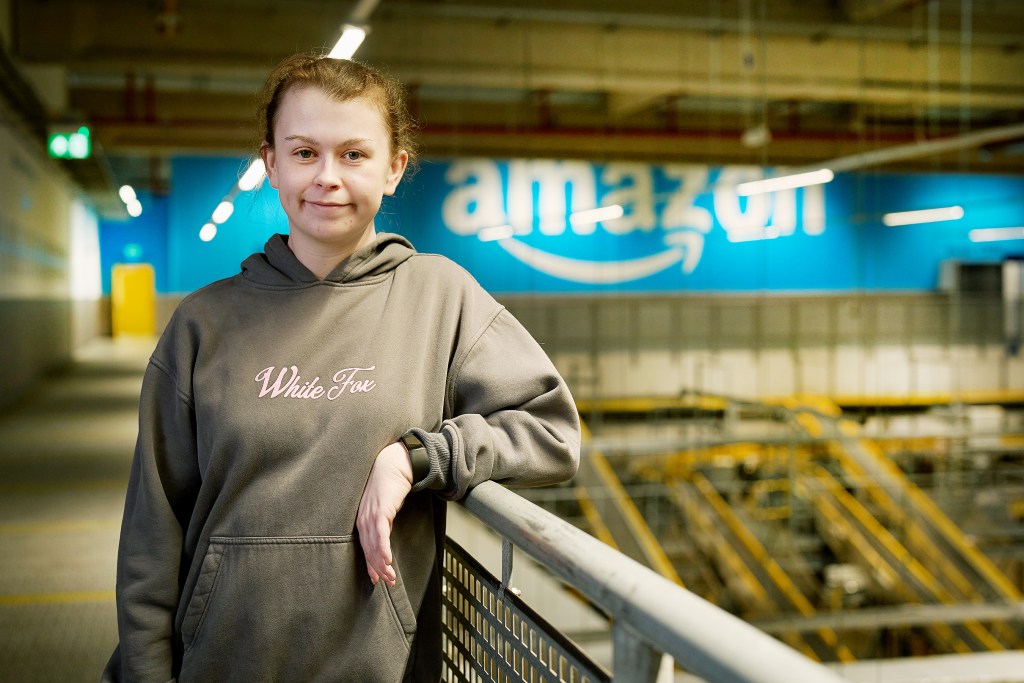 A young woman in a grey “White Fox” hoodie stands inside Amazon’s Swansea fulfilment centre with the Amazon logo behind her.