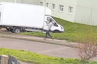 A CCTV screengrab showing a man in a grey top and dark trousers running along a pavement in a residential street, holding an object that appears to be a handgun.