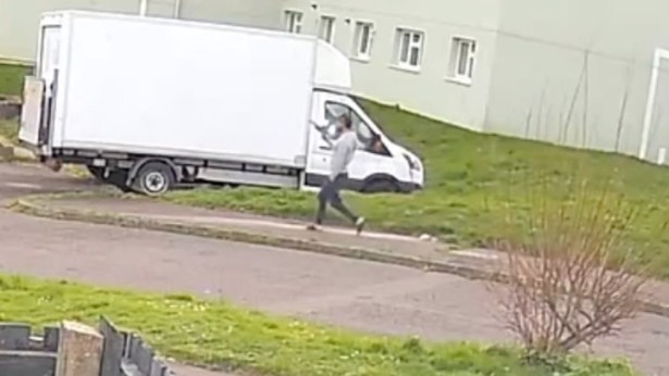 A CCTV screengrab showing a man in a grey top and dark trousers running along a pavement in a residential street, holding an object that appears to be a handgun.