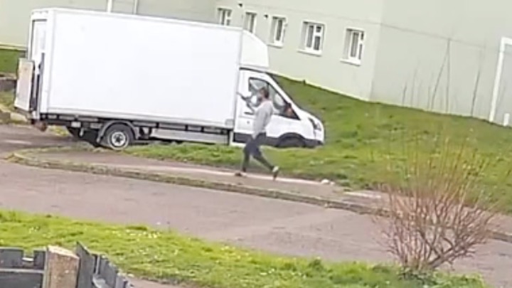 A CCTV screengrab showing a man in a grey top and dark trousers running along a pavement in a residential street, holding an object that appears to be a handgun.