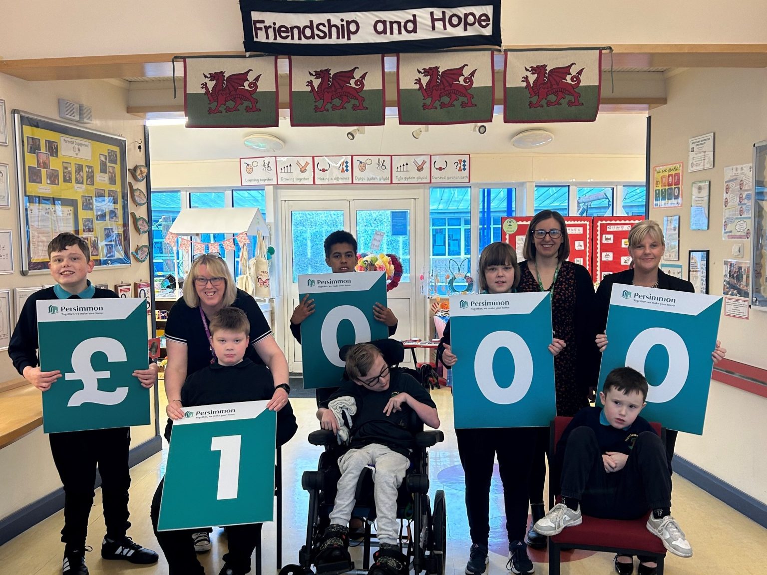 Group of schoolchildren and teachers holding large cheque boards displaying £1,000 inside a school, with Welsh flags and a “Friendship and Hope” banner above.