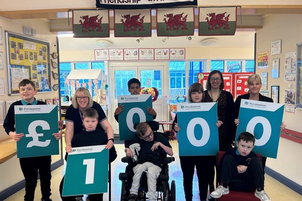 Group of schoolchildren and teachers holding large cheque boards displaying £1,000 inside a school, with Welsh flags and a “Friendship and Hope” banner above.
