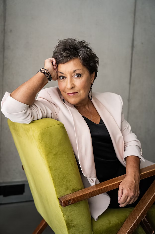 Modern portrait of Ria Jones seated in a green chair, wearing a light pink blazer and looking directly at the camera; the West End performer began her career with Swansea Amateur Operatic Society.