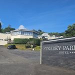 A wide-angle view of the Stradey Park Hotel entrance in Llanelli, showing the hotel's prominent wooden sign with four stars and the main building on the hillside under a clear blue sky.