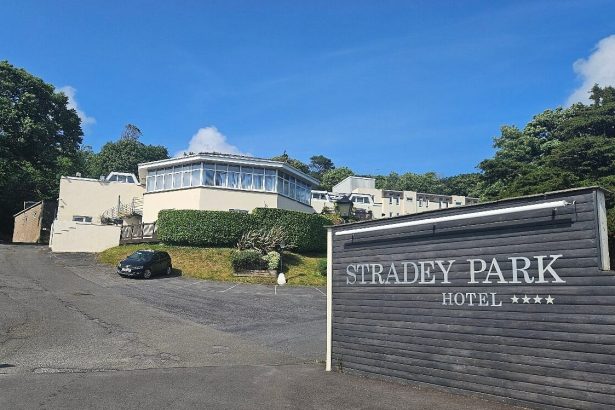A wide-angle view of the Stradey Park Hotel entrance in Llanelli, showing the hotel's prominent wooden sign with four stars and the main building on the hillside under a clear blue sky.
