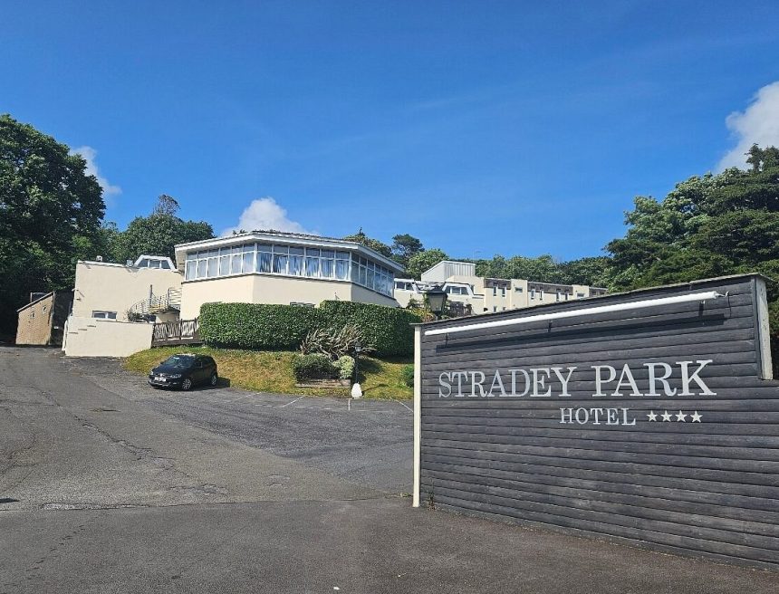 A wide-angle view of the Stradey Park Hotel entrance in Llanelli, showing the hotel's prominent wooden sign with four stars and the main building on the hillside under a clear blue sky.