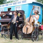 Two street performers with musical instruments performing outdoors beside a colourful caravan and upright piano.