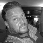 A black and white headshot of David Rogers, a middle-aged man with short, dark, swept-back hair, a short beard, and a light-coloured polo shirt.