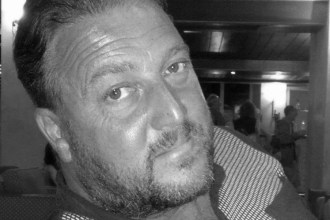 A black and white headshot of David Rogers, a middle-aged man with short, dark, swept-back hair, a short beard, and a light-coloured polo shirt.