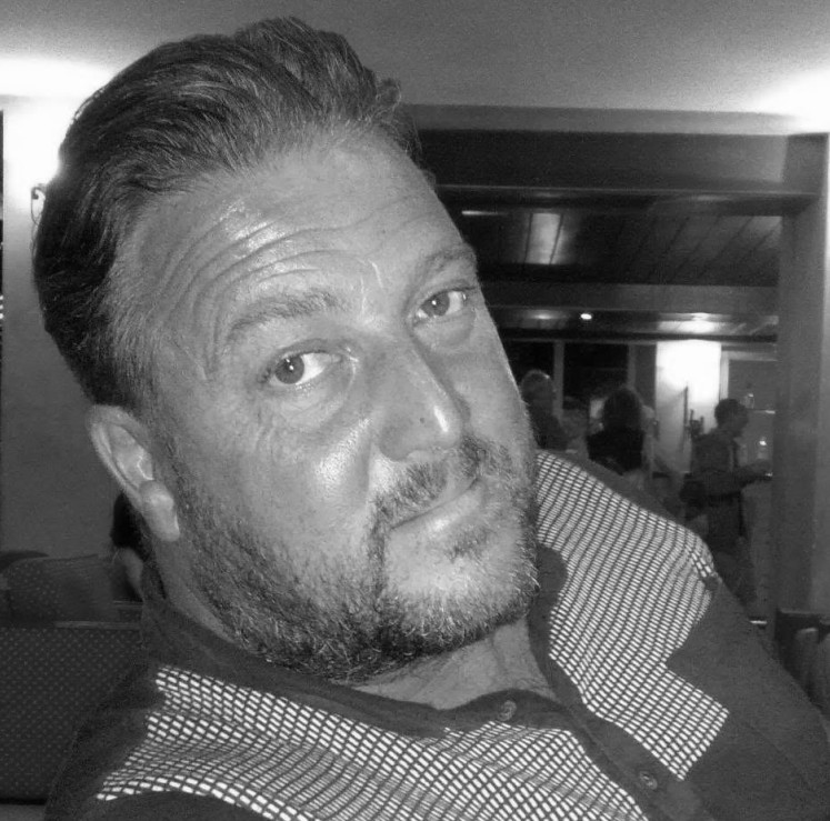 A black and white headshot of David Rogers, a middle-aged man with short, dark, swept-back hair, a short beard, and a light-coloured polo shirt.