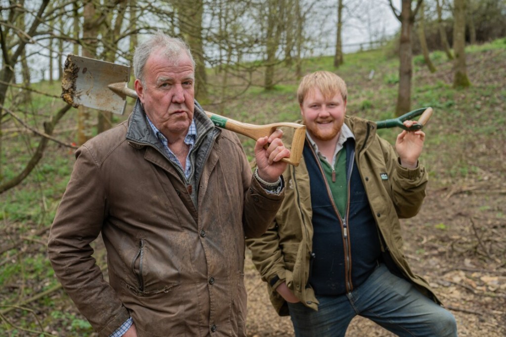 Jeremy Clarkson and Kaleb Cooper hold tools over their shoulders while standing in a wooded farm area.
