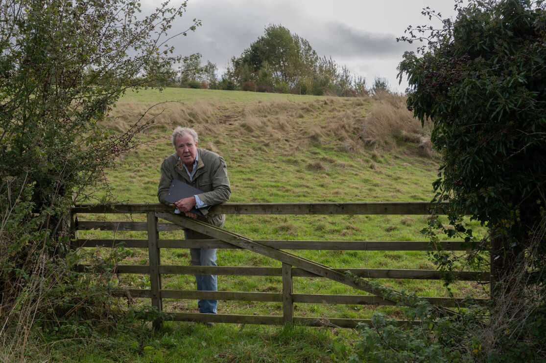 Jeremy Clarkson leans on a wooden gate looking out across a grassy field on his farm.