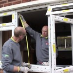 Two men wearing grey ops fitting a uPVC window into a home. One man is holding up a yellow measuring tape.
