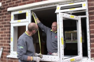 Two men wearing grey ops fitting a uPVC window into a home. One man is holding up a yellow measuring tape.