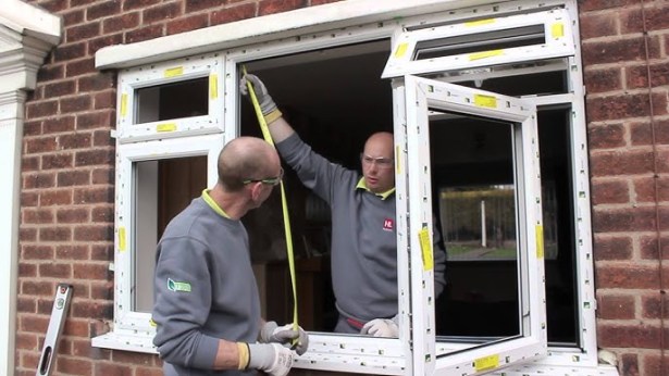 Two men wearing grey ops fitting a uPVC window into a home. One man is holding up a yellow measuring tape.