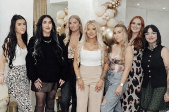 Seven women stand smiling together inside the Dolly Rocks salon, decorated with cream and gold balloon arrangements. Salon owner Jodie Lloyd stands in the centre wearing a white top and beige trousers.
