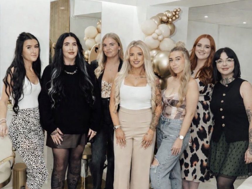 Seven women stand smiling together inside the Dolly Rocks salon, decorated with cream and gold balloon arrangements. Salon owner Jodie Lloyd stands in the centre wearing a white top and beige trousers.