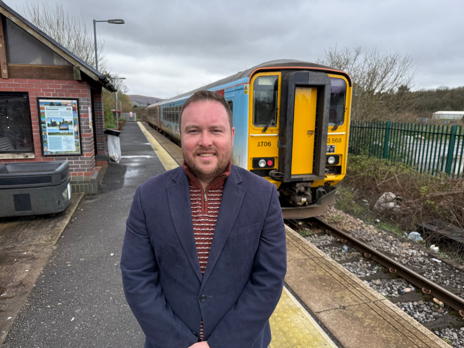Calum Higgins standing on the platform at Ammanford railway station with a yellow and blue train behind him and station signage visible.