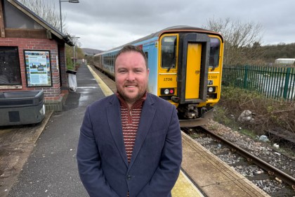 Calum Higgins standing on the platform at Ammanford railway station with a yellow and blue train behind him and station signage visible.