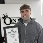 A smiling barber holding a framed certificate reading “2026 Finalist” inside a modern barbershop
