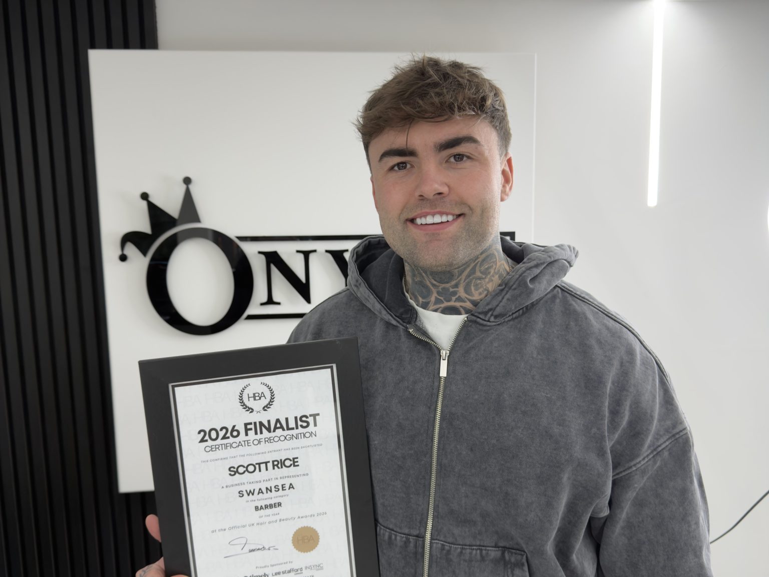 A smiling barber holding a framed certificate reading “2026 Finalist” inside a modern barbershop