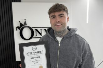 A smiling barber holding a framed certificate reading “2026 Finalist” inside a modern barbershop