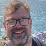 A smiling middle-aged man with reddish-brown hair, a greying beard and black-framed glasses, photographed outdoors with the sea and rocky coastline visible behind him.