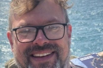 A smiling middle-aged man with reddish-brown hair, a greying beard and black-framed glasses, photographed outdoors with the sea and rocky coastline visible behind him.