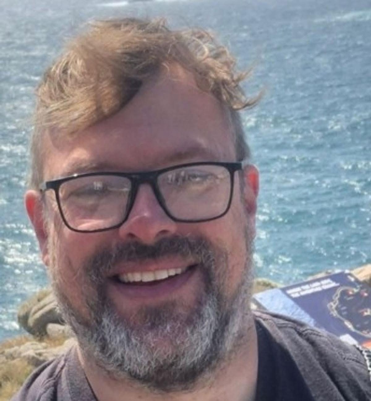 A smiling middle-aged man with reddish-brown hair, a greying beard and black-framed glasses, photographed outdoors with the sea and rocky coastline visible behind him.
