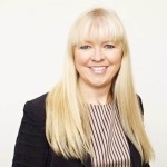 Professional portrait of Marianne Økland, with long blonde hair, wearing a black jacket and striped shirt against a plain white background.