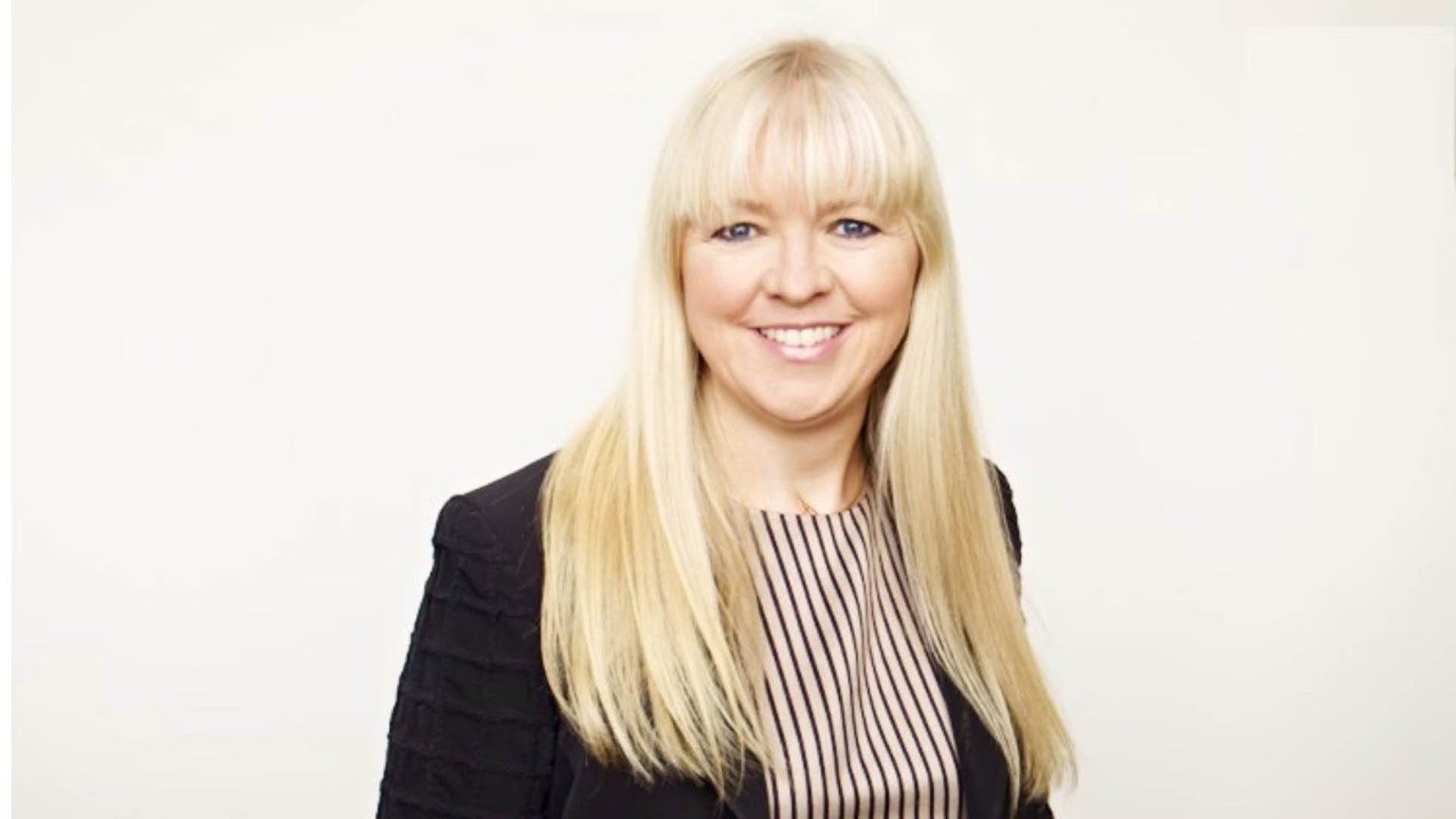 Professional portrait of Marianne Økland, with long blonde hair, wearing a black jacket and striped shirt against a plain white background.
