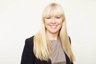 Professional portrait of Marianne Økland, with long blonde hair, wearing a black jacket and striped shirt against a plain white background.