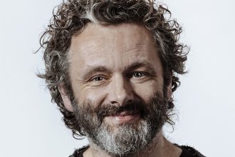 Portrait of Welsh actor Michael Sheen smiling against a plain background.