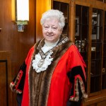 A smiling Penny Matthews wearing the official red and fur-trimmed robes and chain of office of the Deputy Lord Mayor of Swansea, standing in a wood-panelled room.