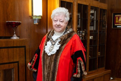A smiling Penny Matthews wearing the official red and fur-trimmed robes and chain of office of the Deputy Lord Mayor of Swansea, standing in a wood-panelled room.