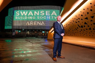 Chief Executive of Swansea Building Society standing outside Swansea Building Society Arena at night with illuminated building signage behind him.